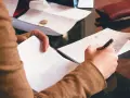 Osoba notuje na kartce, obok leżą dokumenty i monety. Może to być związane z rozliczeniami, np. gwarancja bankowa.