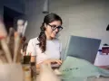Uśmiechnięta kobieta w okularach pracuje przy laptopie, analizując koszty pracodawcy związane z umową zlecenie.