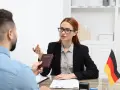Kobieta w okularach przekazuje paszport mężczyźnie, omawiając zarobki w Niemczech. Flaga Niemiec na biurku.