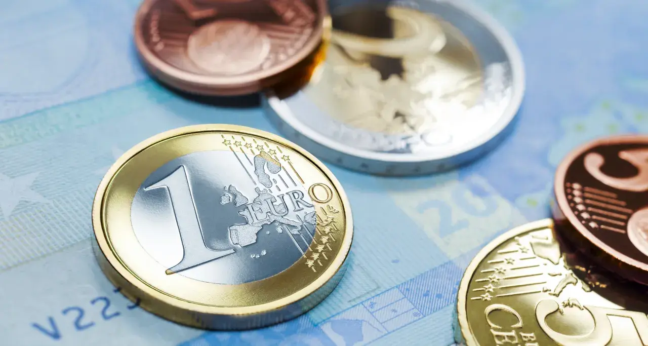 Monety euro, w tym jedna z napisem "EURO" i cyfrą "1", leżą na niebieskim tle banknotu. W tle widać inne monety, miedziane i srebrne, przypominające dawny dinar.