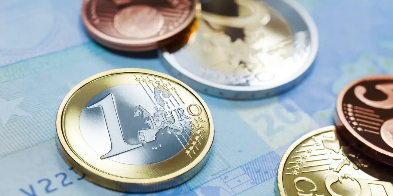 Monety euro, w tym jedna z napisem "EURO" i cyfrą "1", leżą na niebieskim tle banknotu. W tle widać inne monety, miedziane i srebrne, przypominające dawny dinar.
