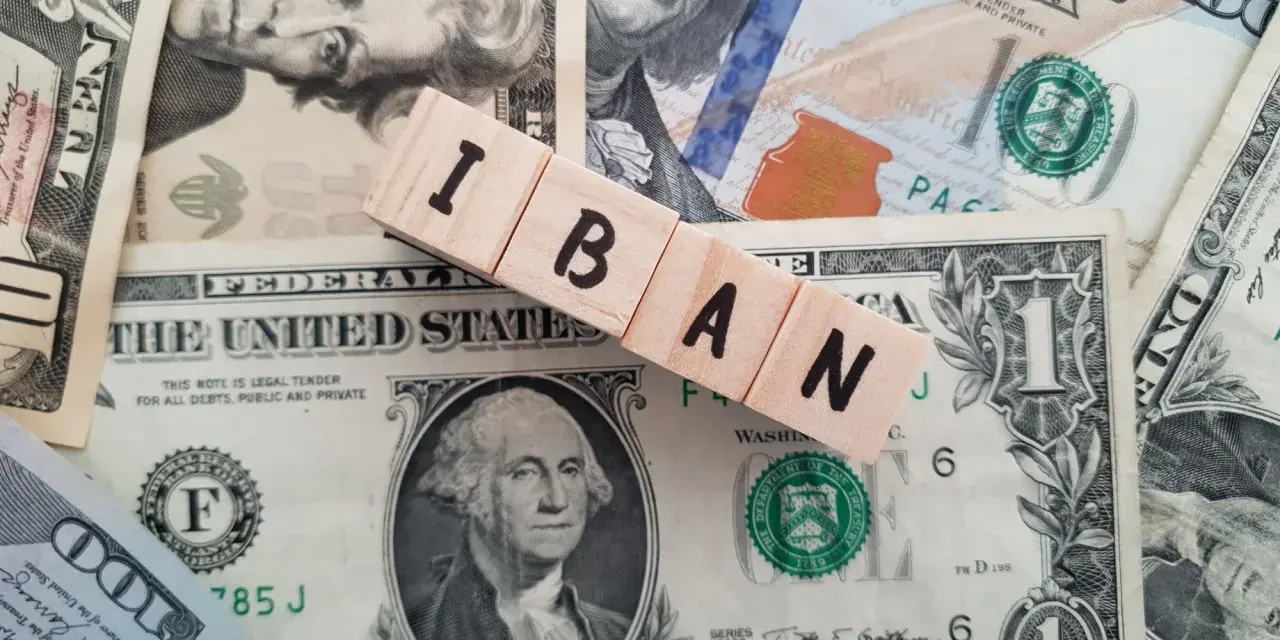 Drewniane klocki z literami IBAN leżą na banknotach dolarowych. Ile cyfr ma numer konta IBAN? To zależy od kraju, ale zazwyczaj jest to 20-34 znaki.