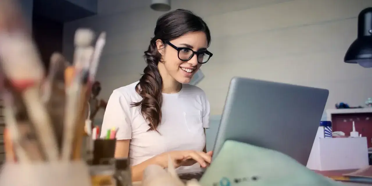 Uśmiechnięta kobieta w okularach pracuje przy laptopie, analizując koszty pracodawcy związane z umową zlecenie.
