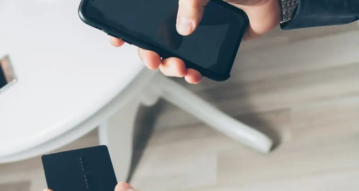Dłoń trzyma smartfon, druga dłoń czarną kartę płatniczą. Szybkie płatności zbliżeniowe, jak w PKO BP, to wygoda.