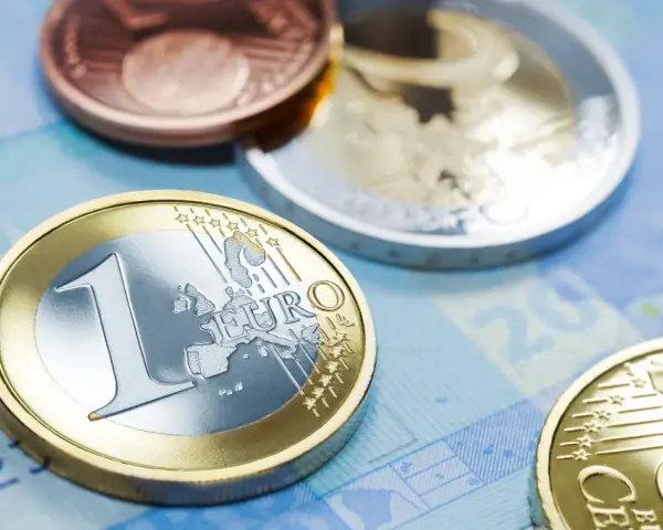 Monety euro, w tym jedna z napisem "EURO" i cyfrą "1", leżą na niebieskim tle banknotu. W tle widać inne monety, miedziane i srebrne, przypominające dawny dinar.
