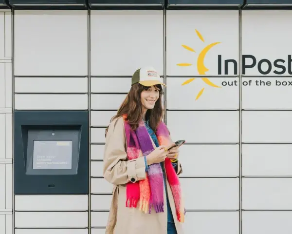 Uśmiechnięta dziewczyna z telefonem w ręku przy paczkomacie InPost. Szybki kurs obsługi, by odebrać przesyłkę.