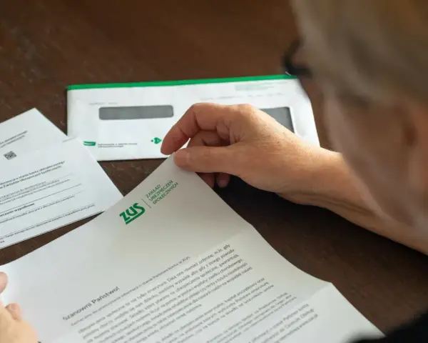 Dłoń starszej osoby przegląda dokumenty z ZUS. W tle koperta z logo ZUS.