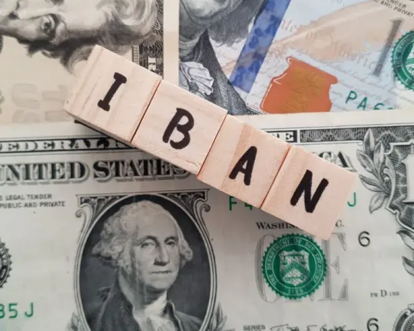 Drewniane klocki z literami IBAN leżą na banknotach dolarowych. Ile cyfr ma numer konta IBAN? To zależy od kraju, ale zazwyczaj jest to 20-34 znaki.