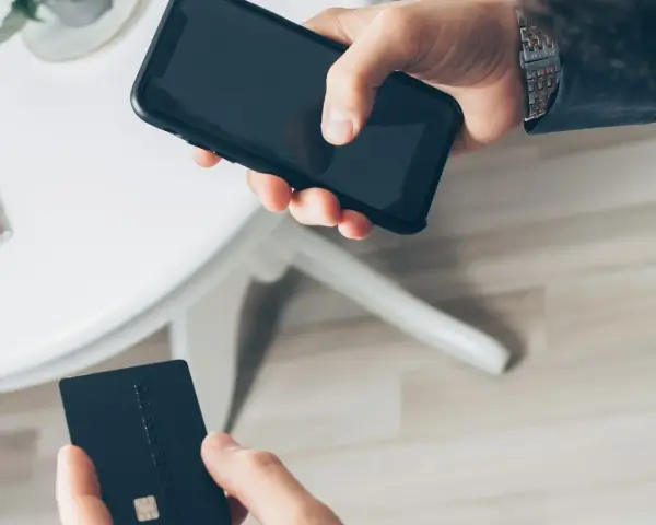 Dłoń trzyma smartfon, druga dłoń czarną kartę płatniczą. Szybkie płatności zbliżeniowe, jak w PKO BP, to wygoda.