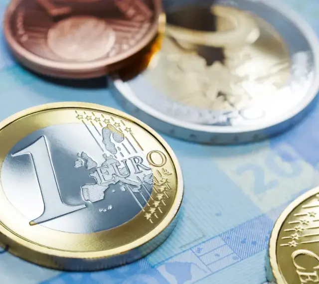 Monety euro, w tym jedna z napisem "EURO" i cyfrą "1", leżą na niebieskim tle banknotu. W tle widać inne monety, miedziane i srebrne, przypominające dawny dinar.