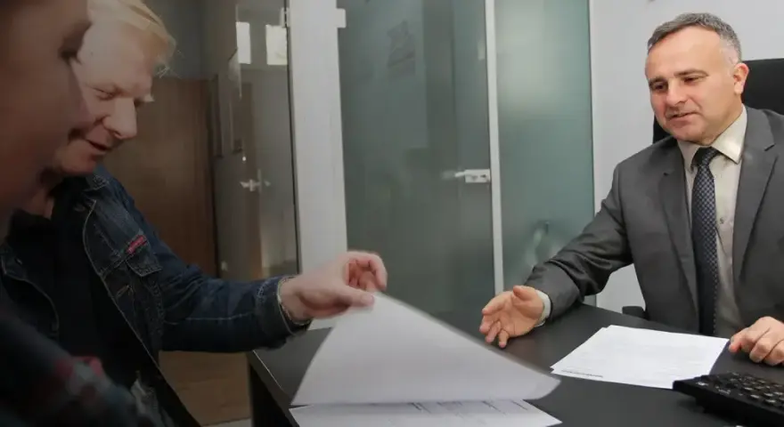 Dwóch mężczyzn przy biurku przegląda dokumenty, jeden w garniturze, drugi w dżinsowej kurtce.