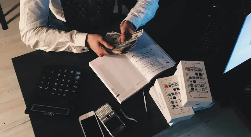 Mężczyzna liczy pieniądze przy biurku z kalkulatorem, notatnikiem, telefonami i pudełkami po iPhone'ach. To może być jego sposób na zarządzanie finansami, być może w kontekście aml.