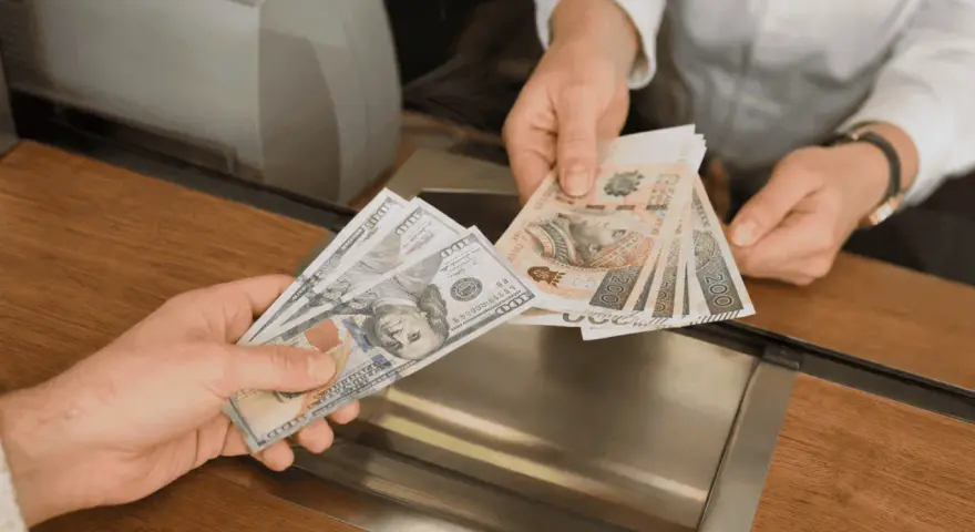 Dwie ręce wymieniają banknoty. Jedna trzyma banknoty 100 dolarów, druga polskie 200 złotych.