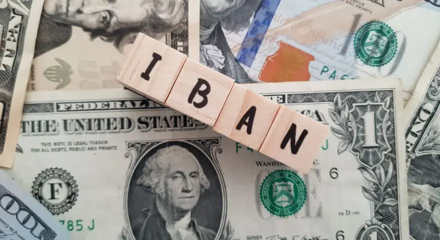 Drewniane klocki z literami IBAN leżą na banknotach dolarowych. Ile cyfr ma numer konta IBAN? To zależy od kraju, ale zazwyczaj jest to 20-34 znaki.