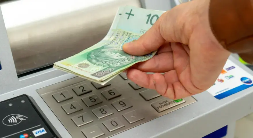 Dłoń wkłada banknoty 100 zł do bankomatu. Zastanawiasz się, ile można wpłacić na konto?
