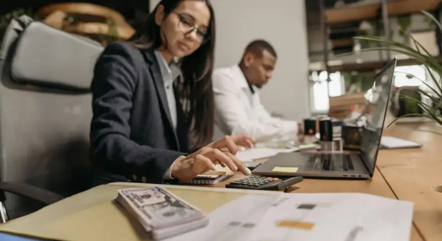 Kobieta w okularach liczy na kalkulatorze, obok stos banknotów i laptop.
