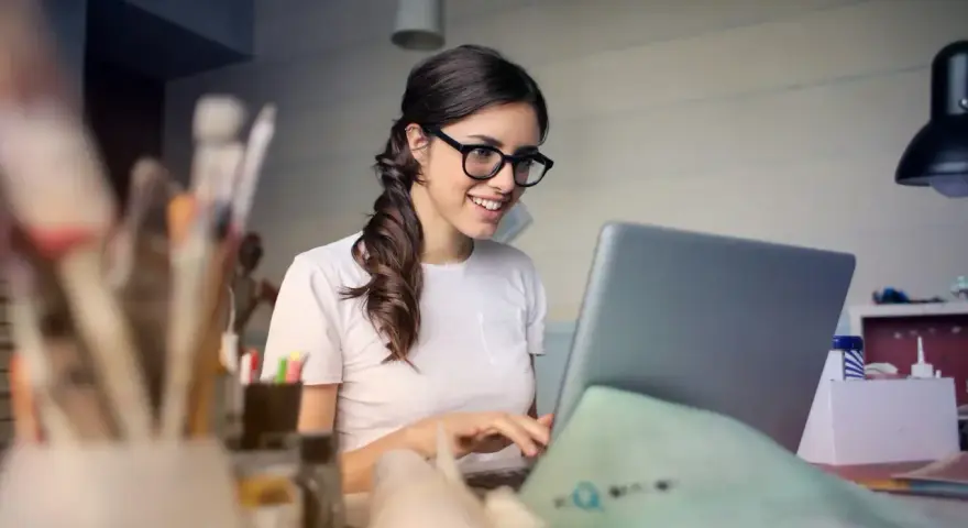Uśmiechnięta kobieta w okularach pracuje przy laptopie, analizując koszty pracodawcy związane z umową zlecenie.
