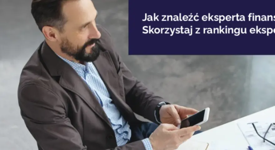 Mężczyzna z brodą trzyma smartfon, siedząc przy biurku z dokumentami.