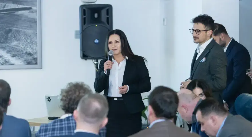 Kobieta w garniturze przemawia do grupy ludzi, obok mężczyzna w okularach.