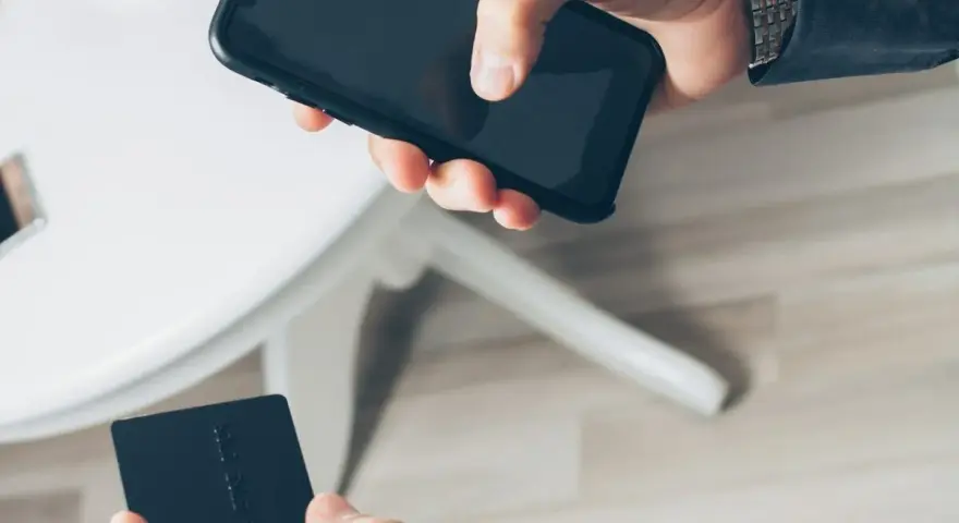 Dłoń trzyma smartfon, druga dłoń czarną kartę płatniczą. Szybkie płatności zbliżeniowe, jak w PKO BP, to wygoda.