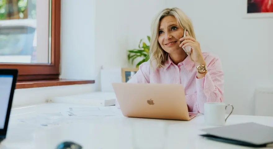 Kobieta w różowej koszuli rozmawia przez telefon, siedząc przy biurku z laptopem.