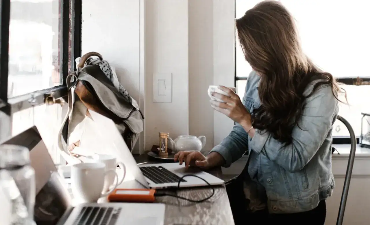 Kobieta pracuje na laptopie, pijąc kawę. To obrazek pokazujący, jak wygląda praca freelancera.